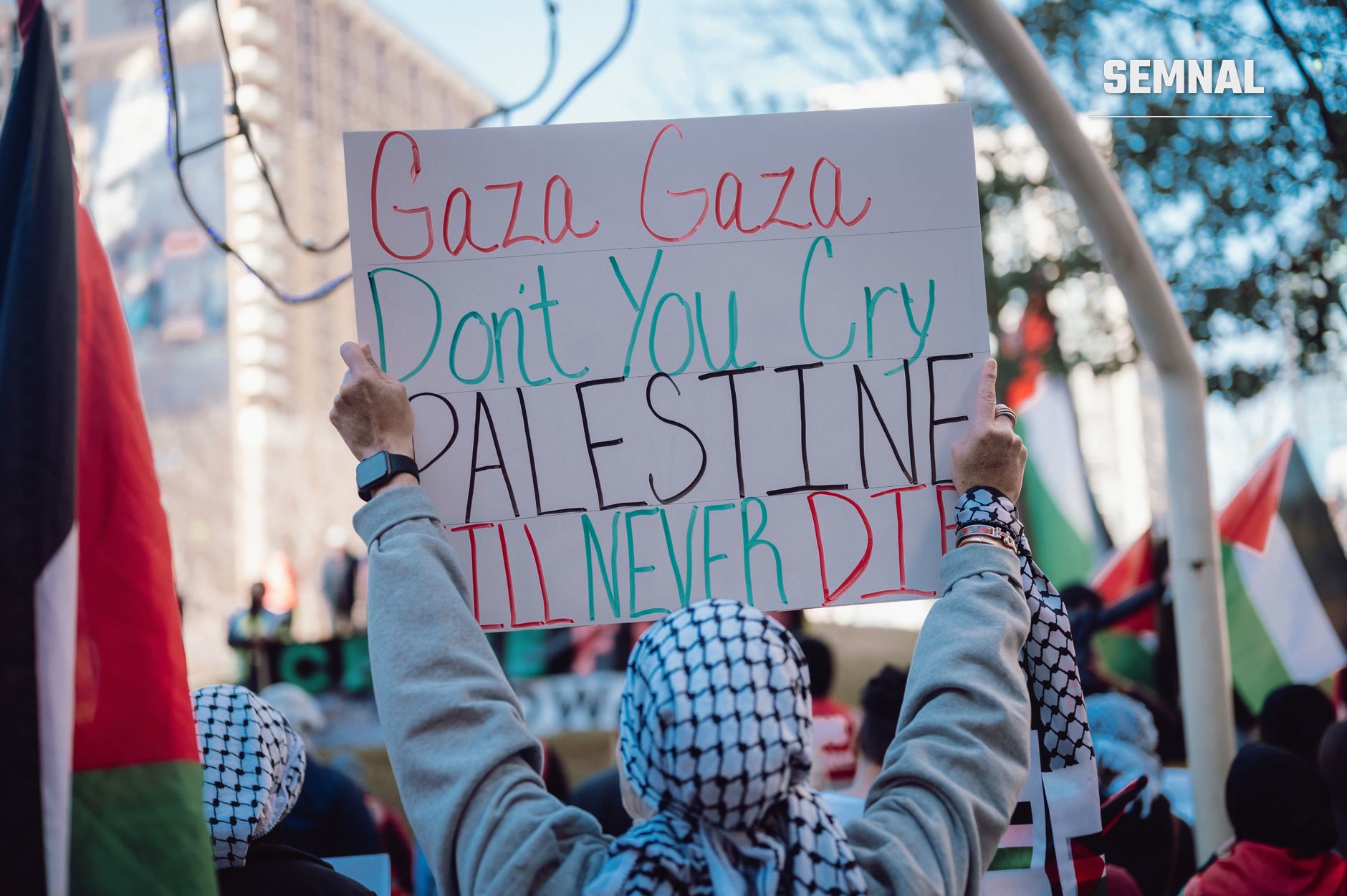 Manifestant la protest care ține o pancartă cu mesaj de susținere pentru Palestina, cu steaguri palestiniene și israeliene în fundal, la o adunare urbană.