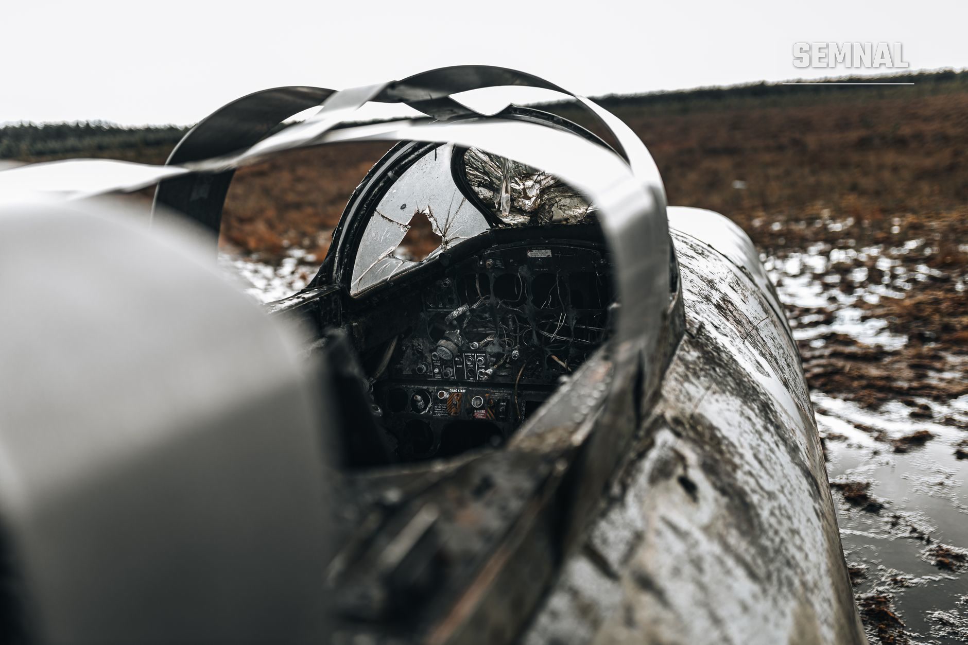 Epavă de avion militar cu fuselaj alb și negru, oglindă ruptă și cockpit distrus pe teren neigienic cu zăpadă.