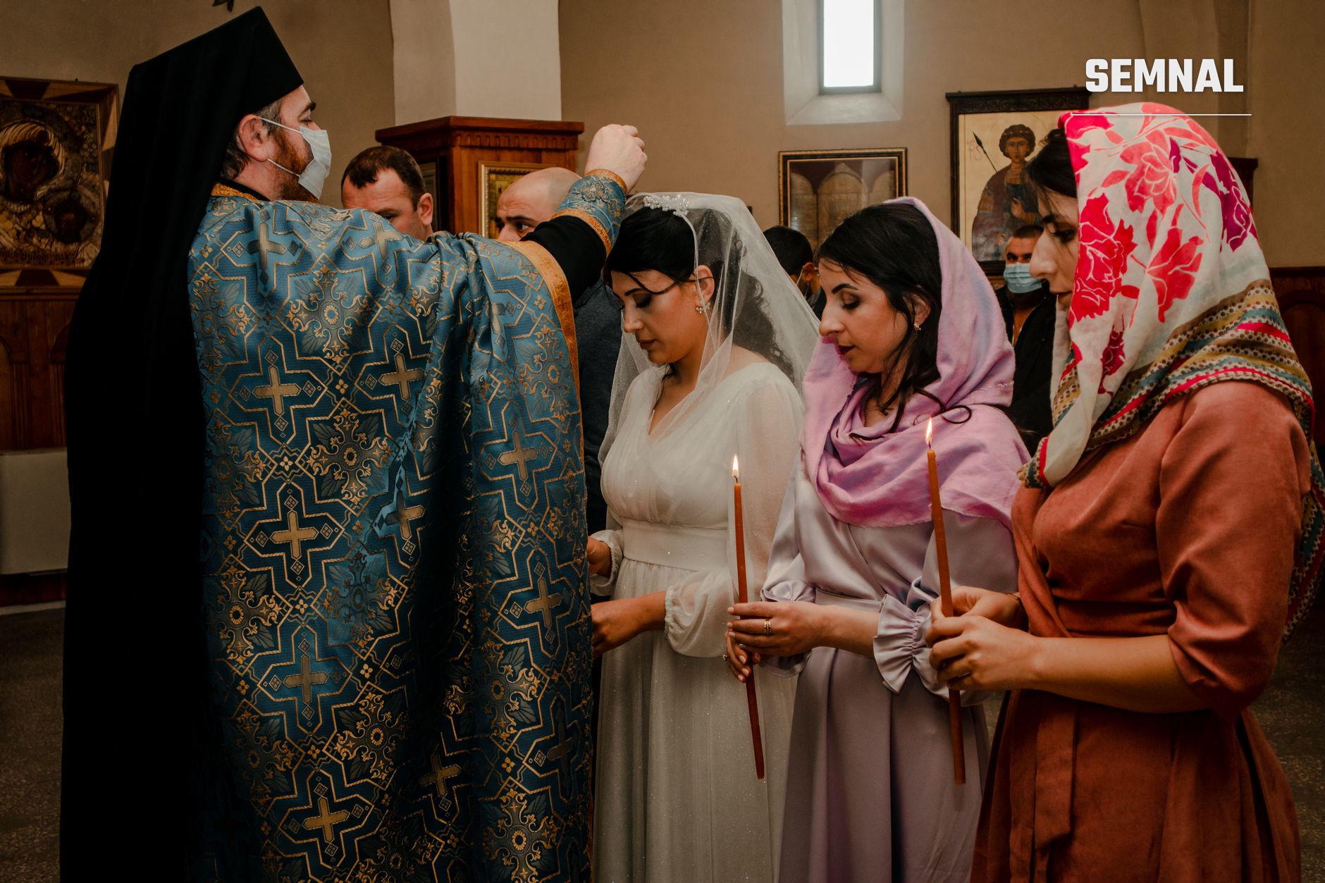 Preot ortodox cu vestminte albastru-aurii oferind binecuvântare unor femei credincioase cu capul acoperit în interiorul unei biserici, ilustrând tradiții și practici religioase românești contemporane.