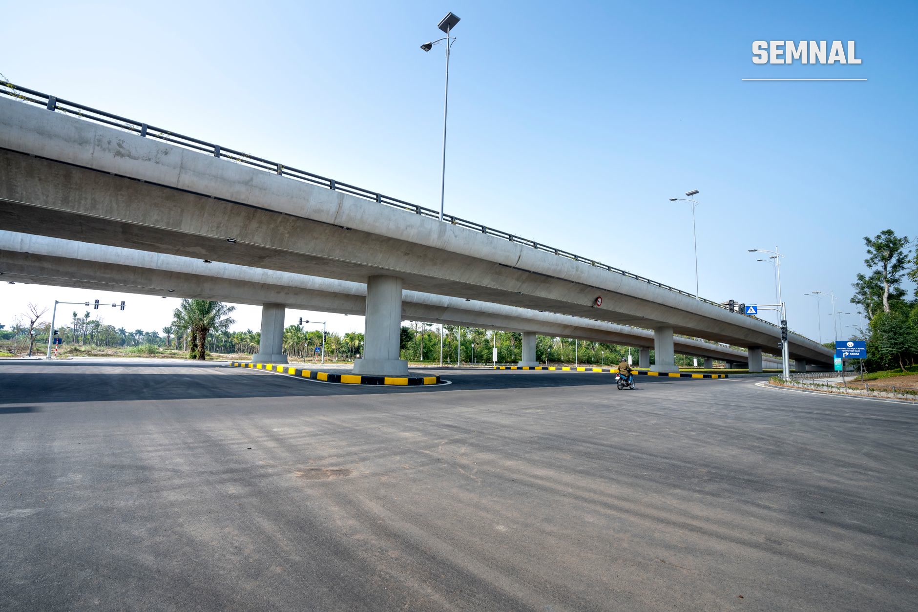 Autostradă modernă cu viaduct din beton și marcare galben-neagră, iluminare solară și semn rutier albastru pe o cărări goală sub cer senin, ilustrând infrastructura de transport rutier actuală.