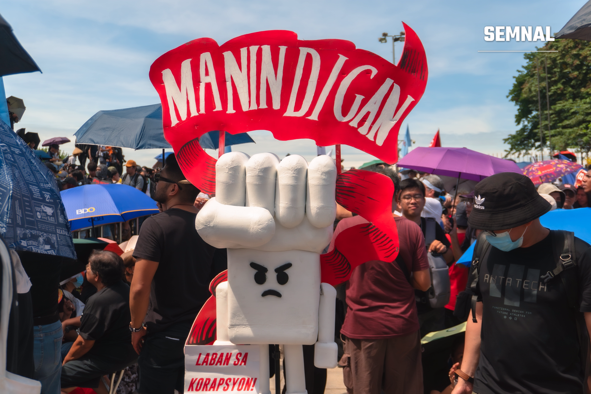 Mascot cu pumn ridicat și expresie furioasă purtând banderolă roșie, în mijlocul unei manifestații împotriva corupției la Jakarta, cu mulțimea de protestatari în fundal.