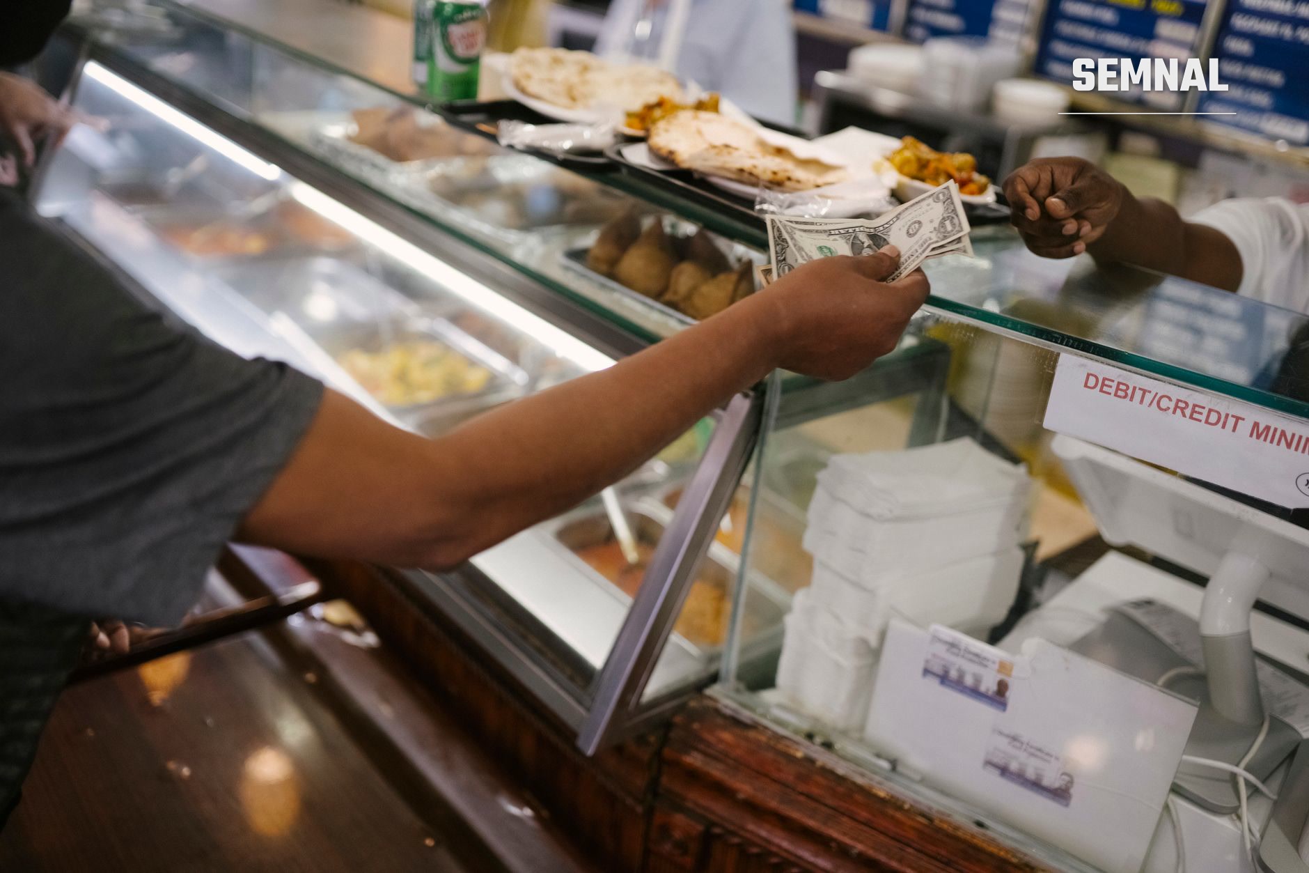 Mână care oferă o bancnotă la tejghel de fast-food, cu vitrină cu mâncare și indicator de plată cu card în fundal