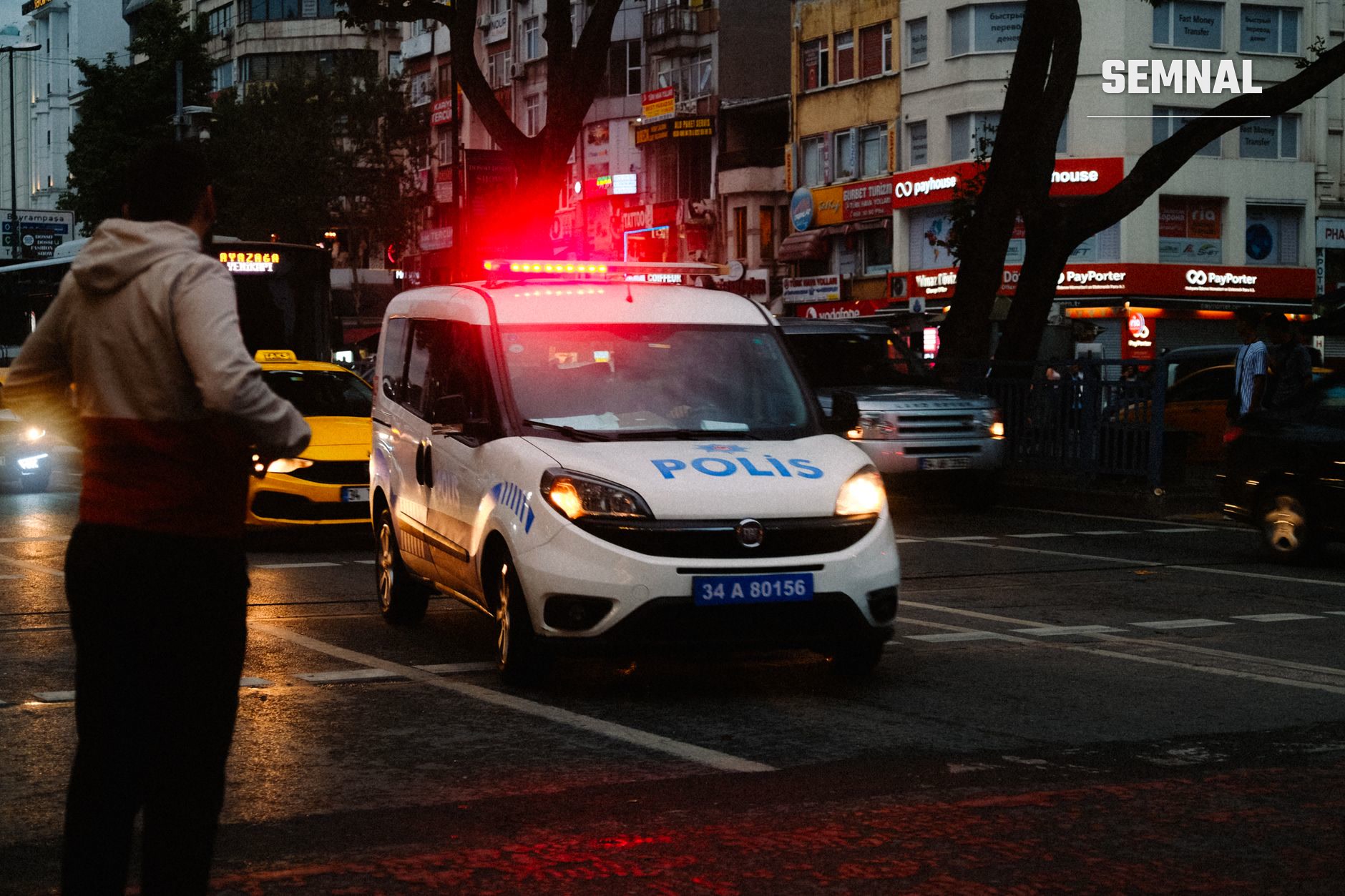 Mașină de poliție albă cu sirene roșii activate în centrul orașului Minneapolis noaptea, în trafic dens cu taxiuri și clădiri iluminate