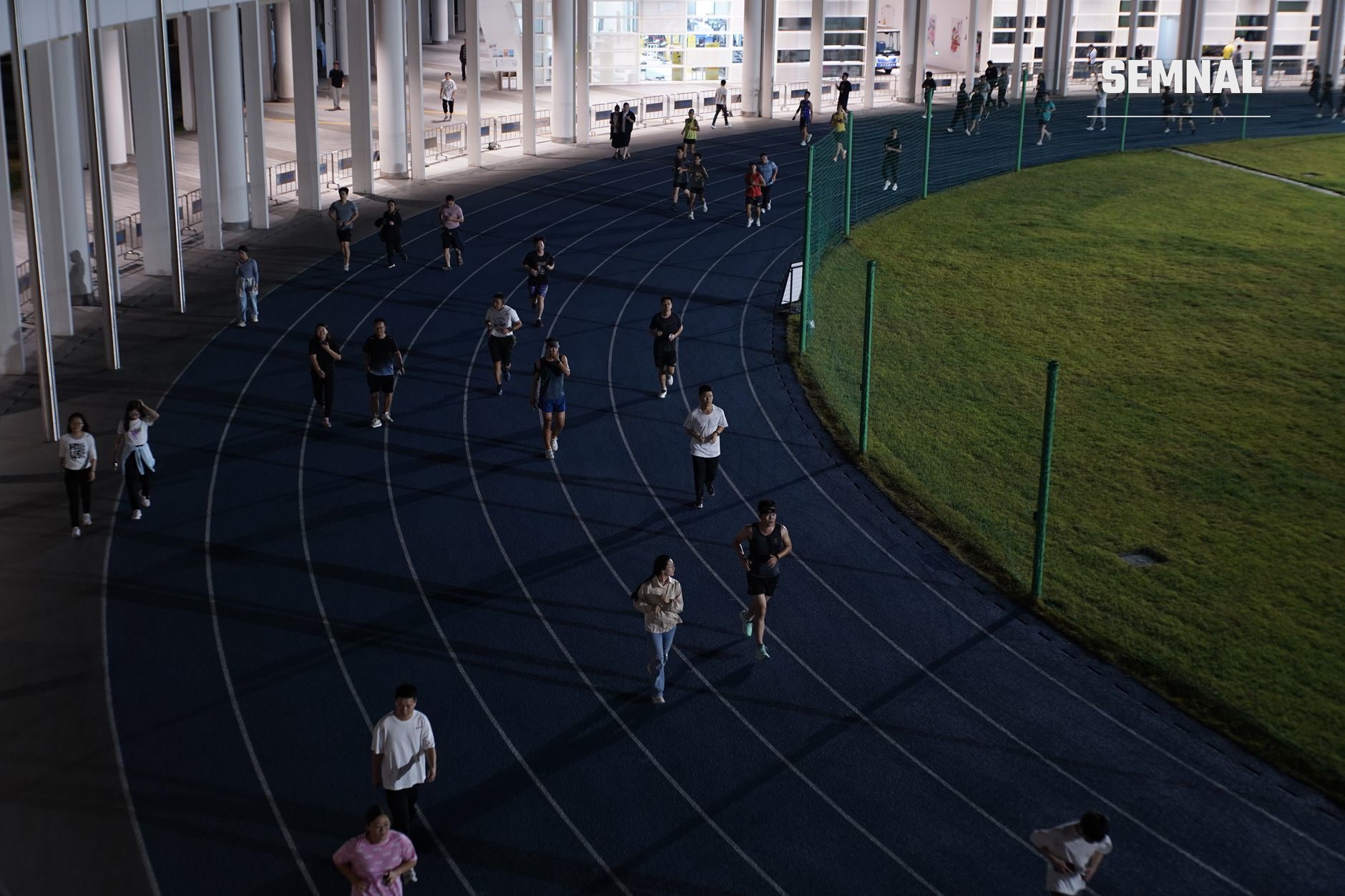 Zeci de alergători distribuiți pe pista de atletism curbă, sub structură de beton, alergând și antrenând în grup pe benzi paralele marcate, evidențiind comunitatea și cursele populare