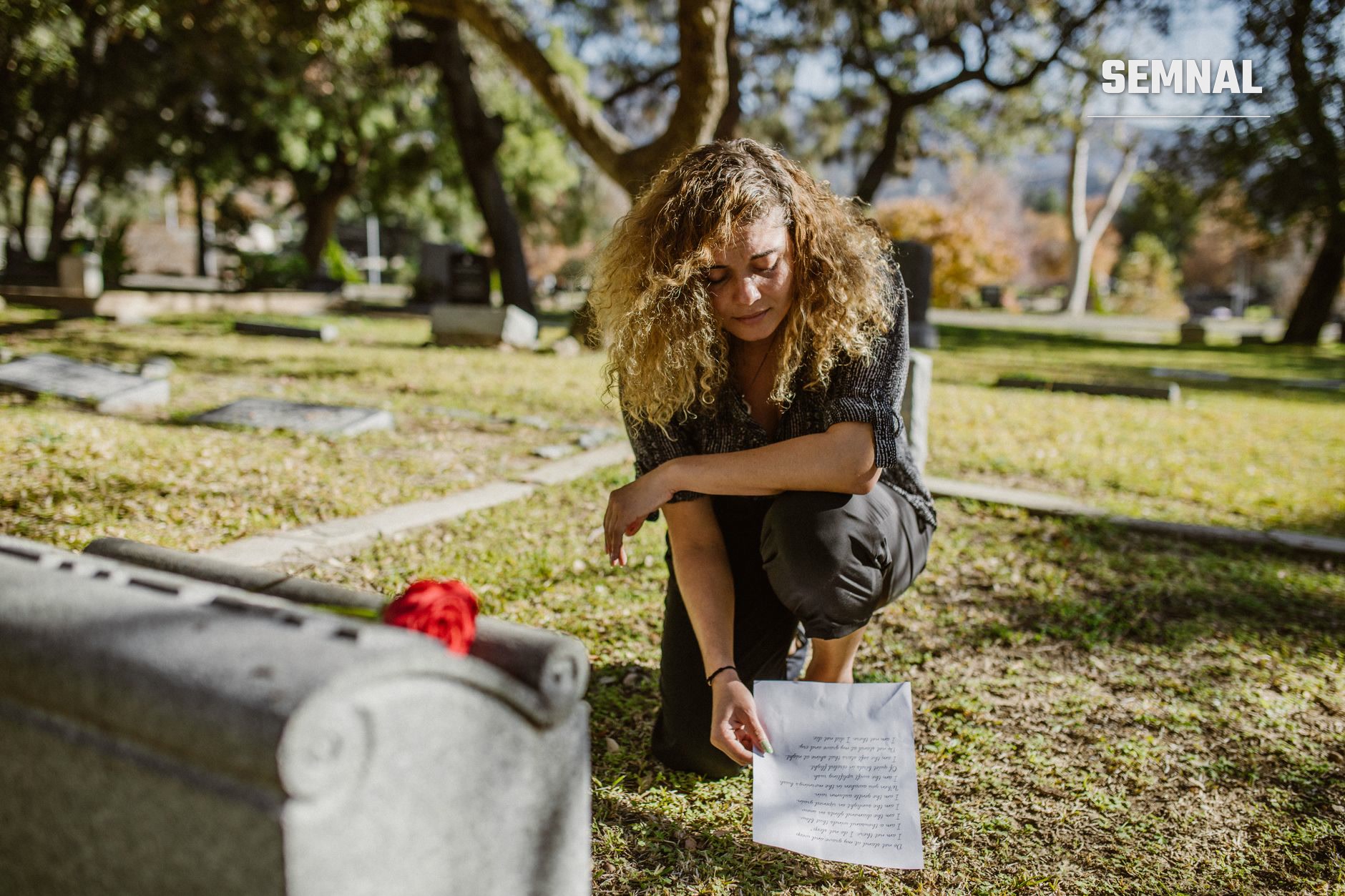 Femeie cu părul creț ținând o scrisoare deasupra mormântului cu flori roșii în cimitir, într-un moment de rugăciune și doliu