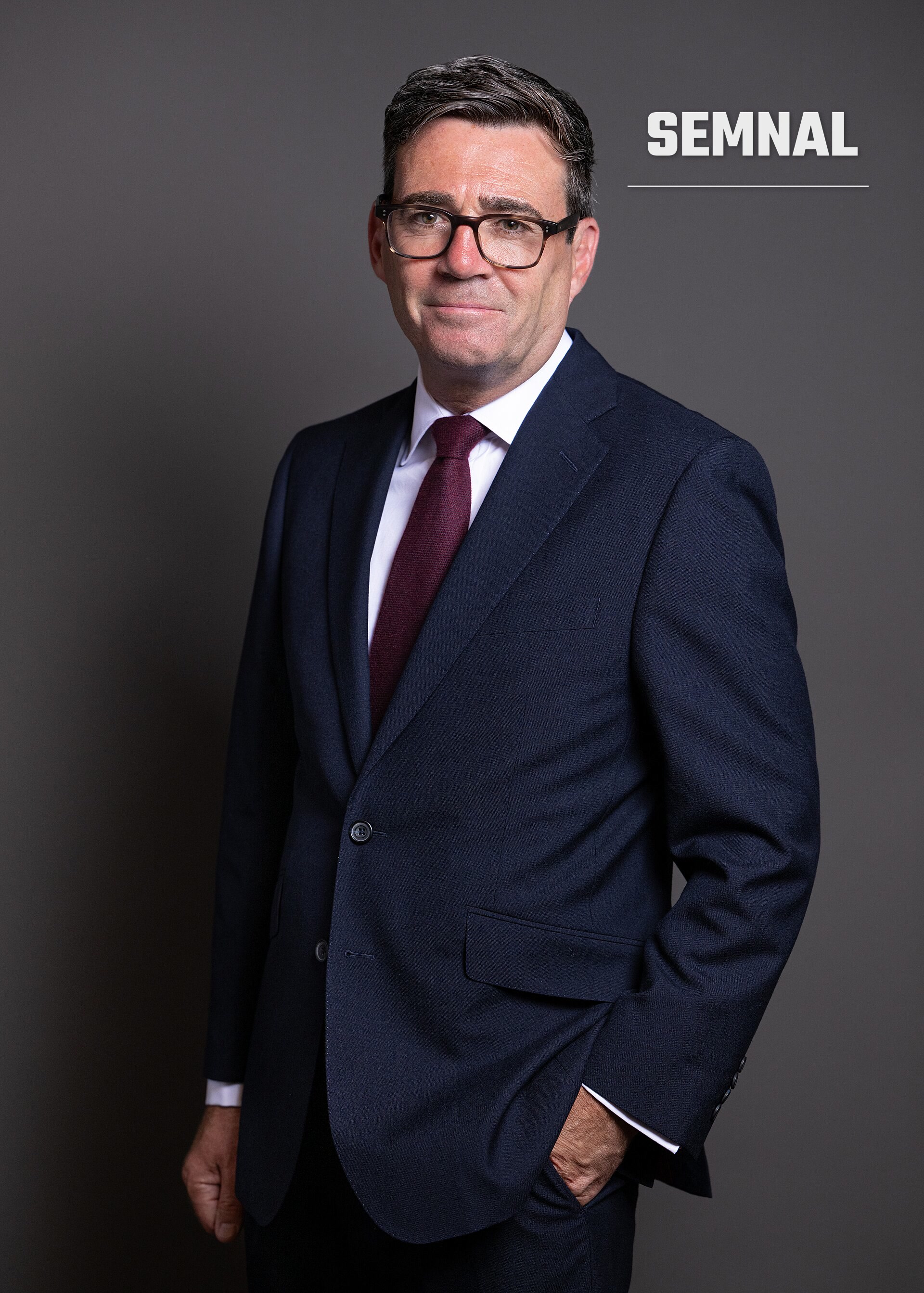 Andy Burnham, portret oficial de presa, barbat cu ochelari in costum bleumarin si cravata bordeaux, pe fundal gri. (Foto: House of Commons)