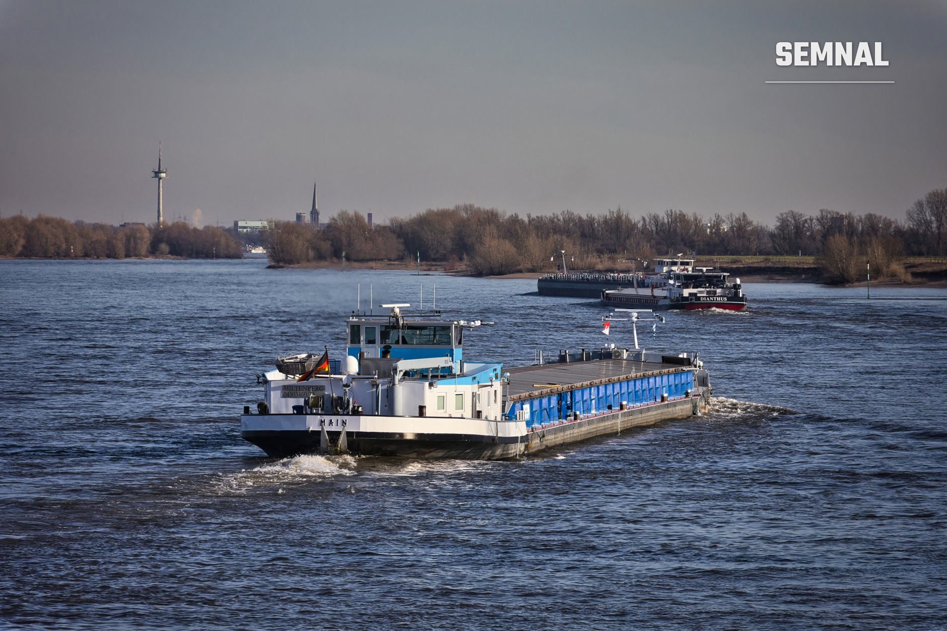Barcă de transport cu container albastru navigând pe fluviu, alături de alte nave și structuri portuare, ilustrând traficul fluvial activ pe canalele de navigație. (Foto: Wolfgang Vrede / Pexels)