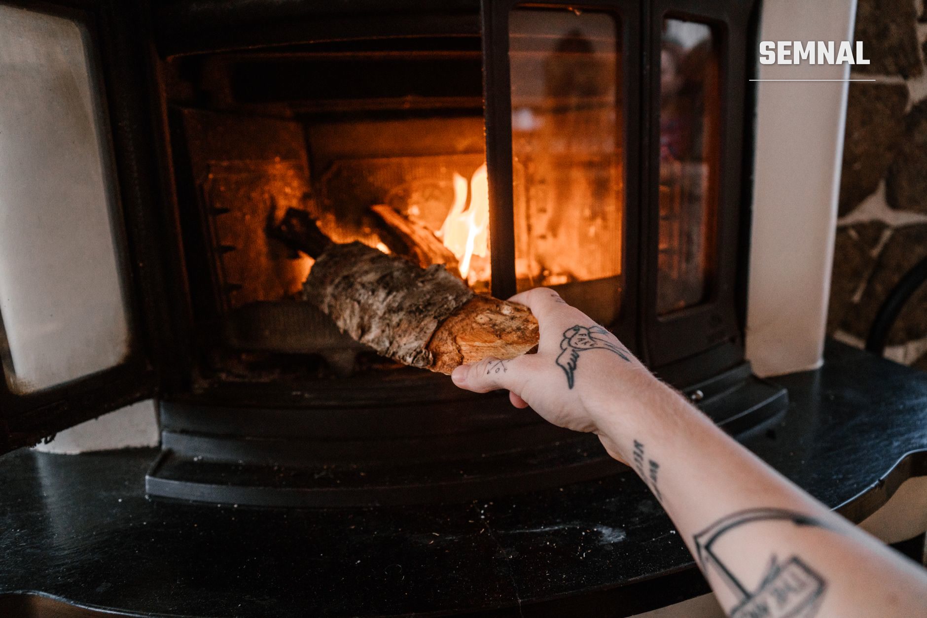 O persoană cu tatuaje pune lemne într-o sobă aprinsă, cu ușa din sticlă deschisă. (Foto: ArtHouse Studio / Pexels)