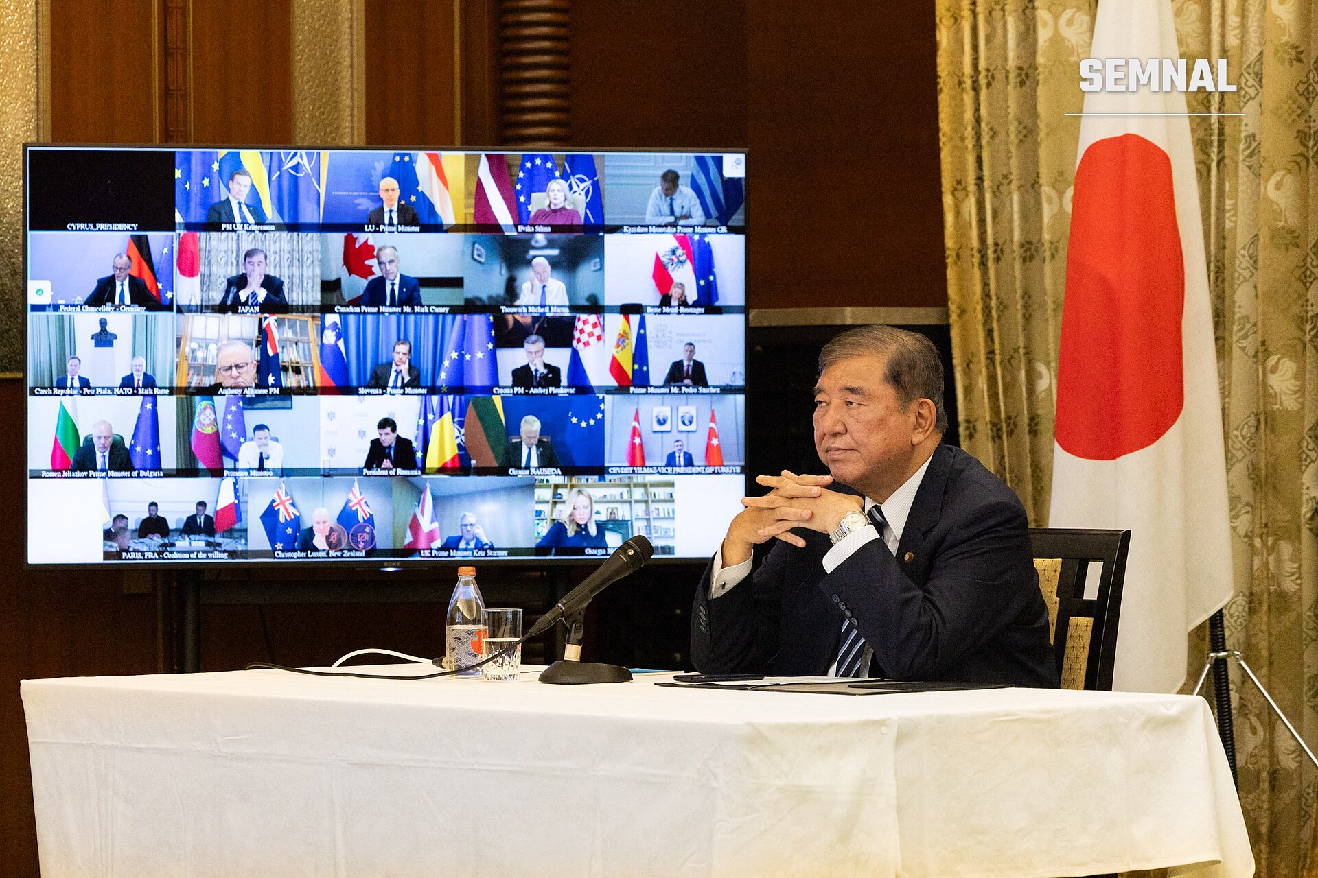 Nicușor Dan, președintele României, seated la o conferință internațională cu ecran video în fundal arătând participanți din mai multe țări, steag japonez vizibil pe dreapta. (Foto: Cabinet Public Affairs Office of Japan)