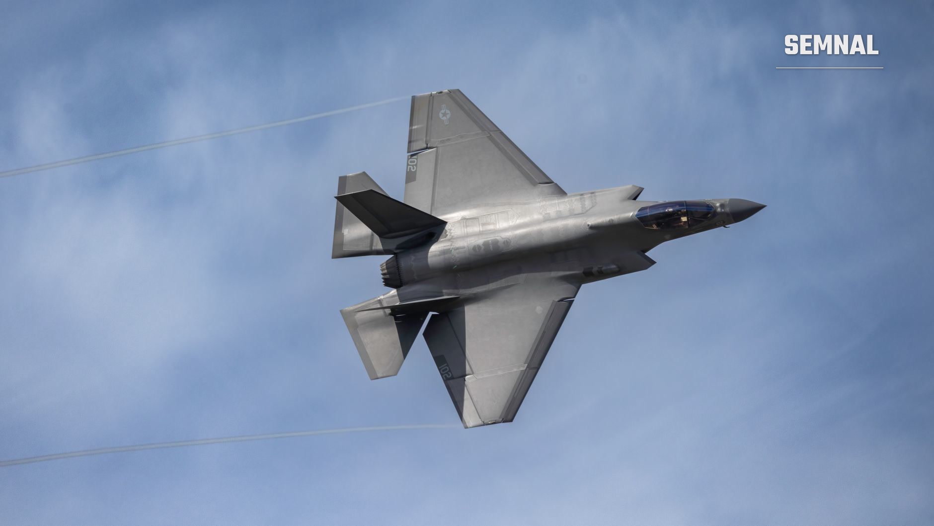 Avion de vânătoare F-35 Lightning II în zbor, vedere de jos pe cer albastru cu nori ușori, având fuzelaj argintiu și aripi delta caracteristice. (Foto: Guy Seela / Pexels)