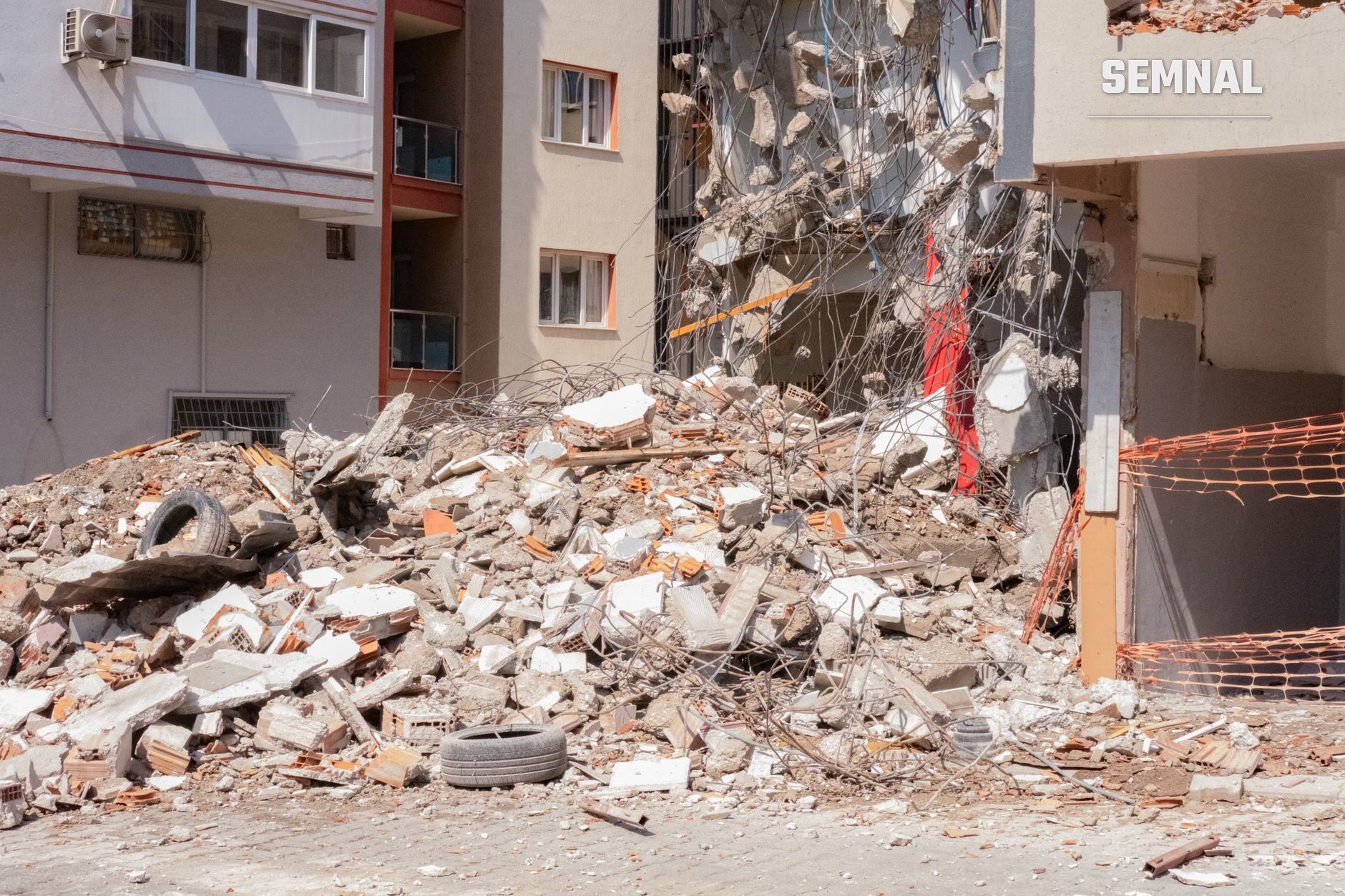 Clădire rezidențială complet distrusă, acoperită cu moloz de beton și dărâmături, cu clădiri vecine avariate și plasă de siguranță roșie pe fațada unei structuri dăunate. (Foto: Doğan Alpaslan Demir / Pexels)