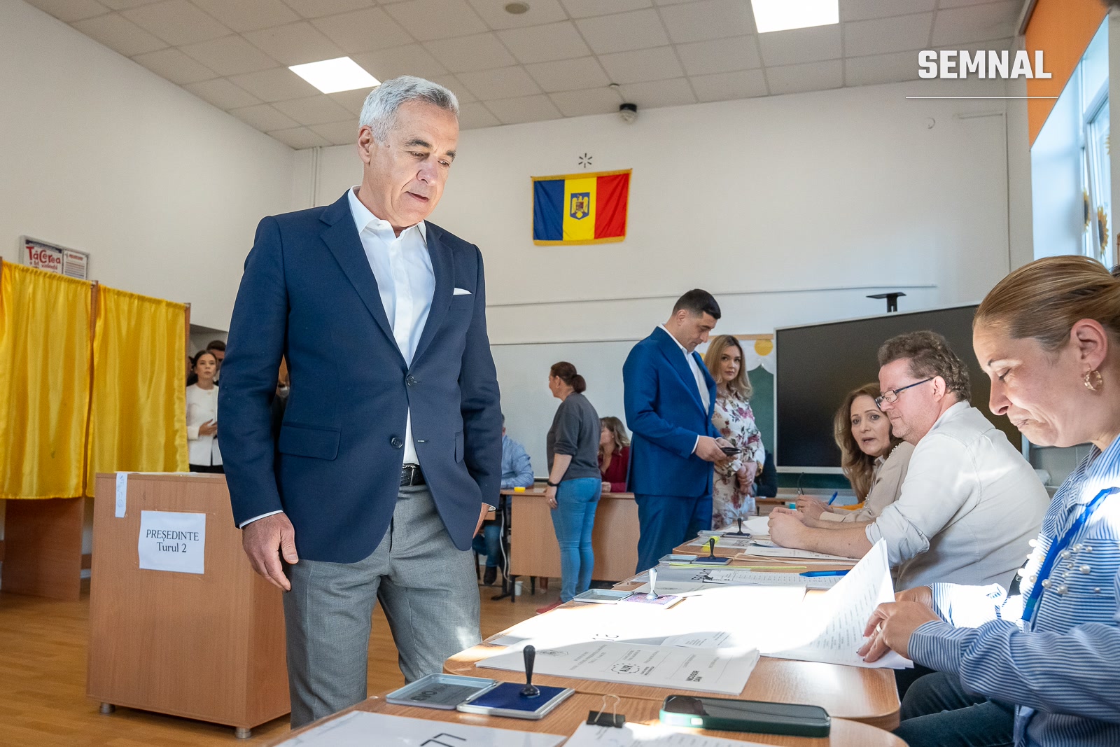Călin Georgescu, îmbrăcat în costum bleumarin și cămașă albă, la lângă o urnă de vot din lemn cu inscripția "PREȘEDINTE Turul 2", într-o sală de vot cu steagul României vizibil pe perete. (Foto: AUR Alianța pentru Unirea Românilor)