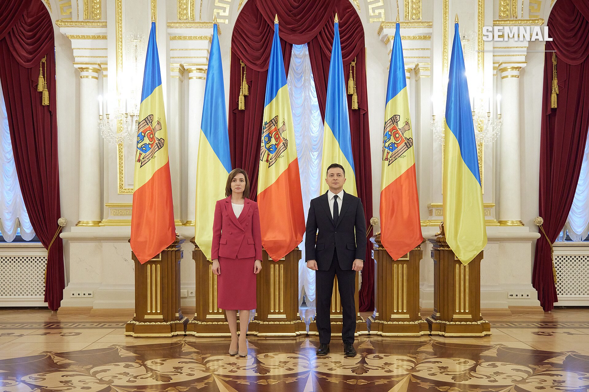 Maia Sandu, președinta Moldovei, într-o sală oficială cu steaguri tricolore, purtând un costum bordo, alături de un oficial, în fața unui decor formal cu perdele roșii și coloane aurii. (Foto: President office of Ukraine)
