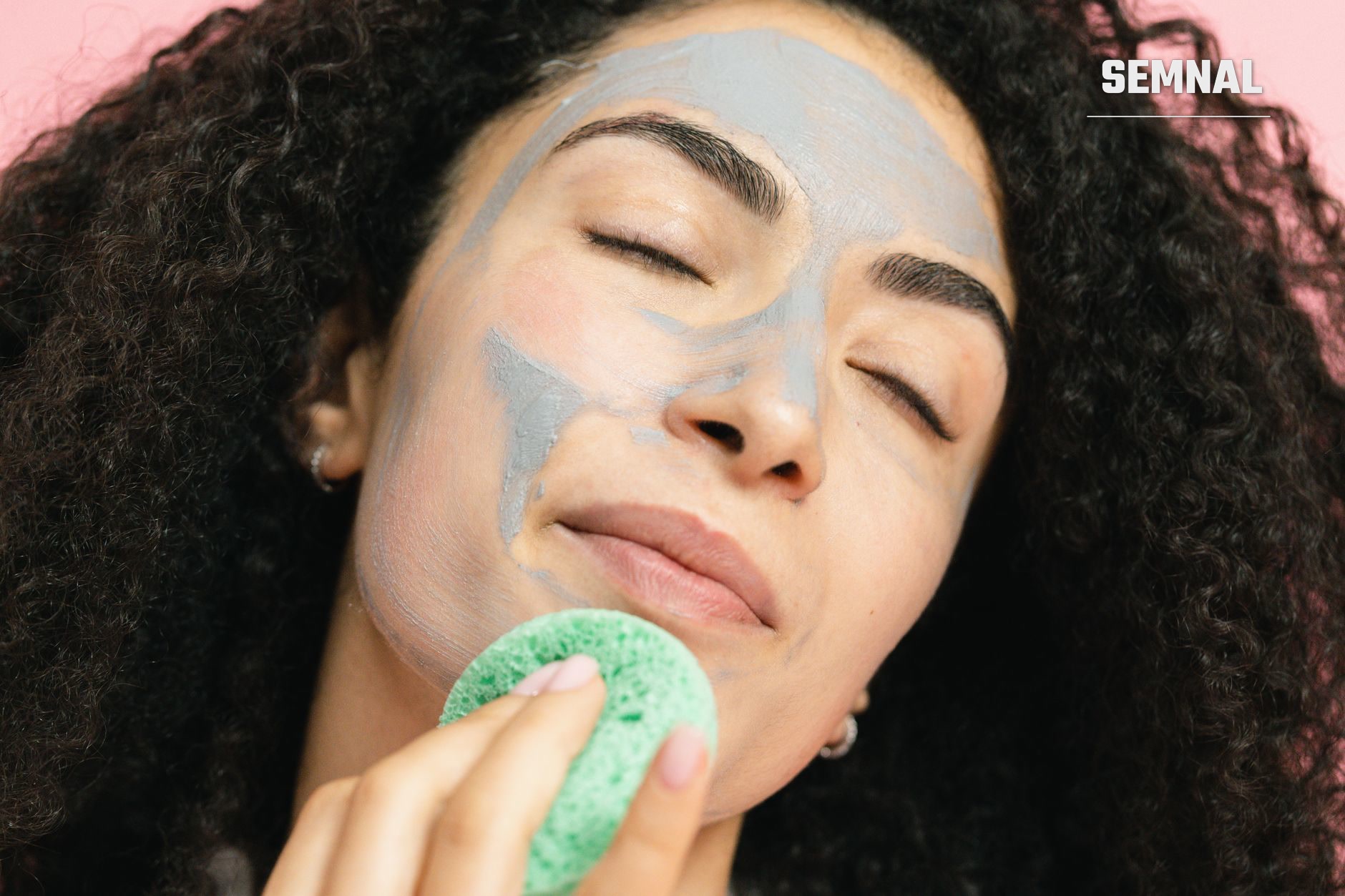 Femeie care își aplică pe față o mască de îngrijire gri-albăstruie cu un burete verde, ca parte a rutinei de îngrijire a pielii (Foto: SHVETS production / Pexels)