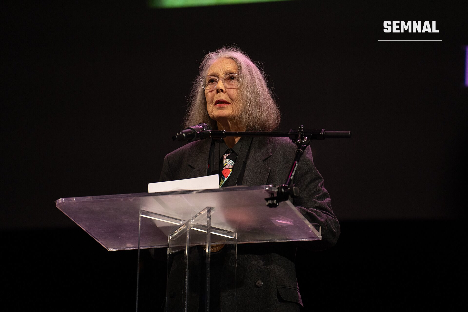 Anne Carson, scriitoare, la o catedră vorbind la o conferință, purtând costum negru și ochelari. (Foto: Jay Dixit)