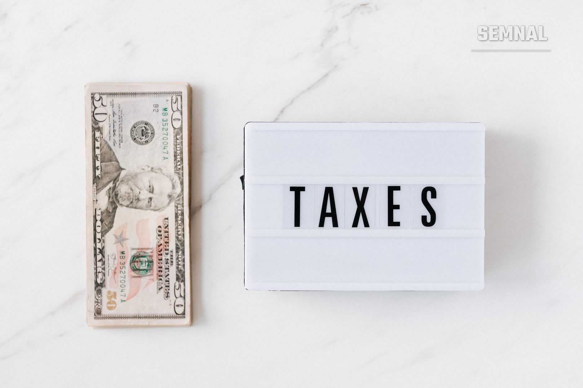 Bancnotă de 50 de dolari americani alături de o lightbox cu cuvântul 'TAXES' pe o suprafață de marmură albă, simbolizând impozitele și paradisurile fiscale (Foto: https://kaboompics.com/ / Pexels)