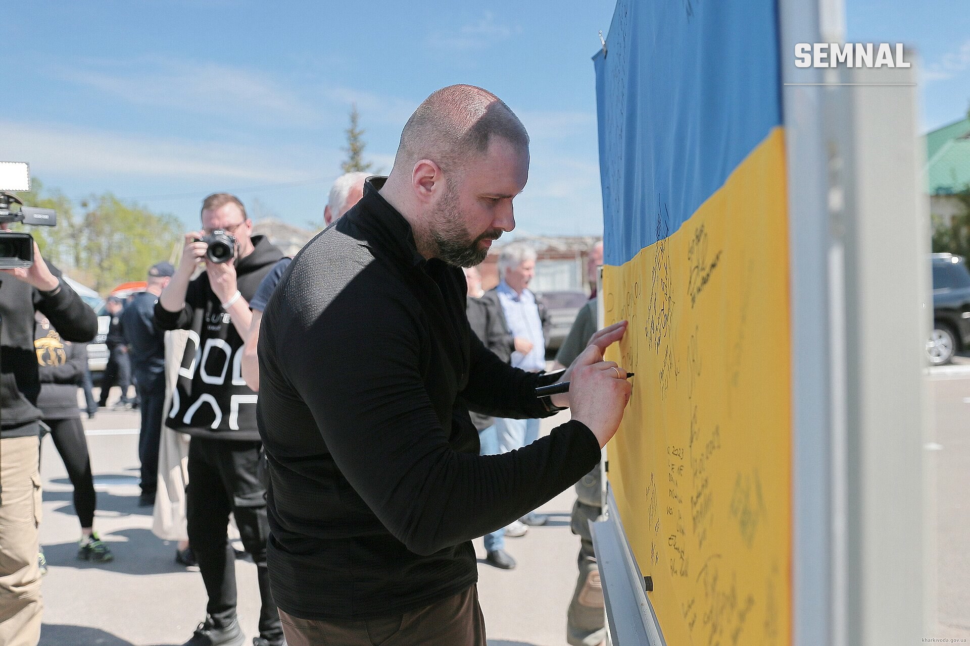 Oleh Syniehubov scrie pe o suprafață galbenă la un eveniment public, purtând jachetă neagră. (Foto: Харківська обласна військова адміністрація)