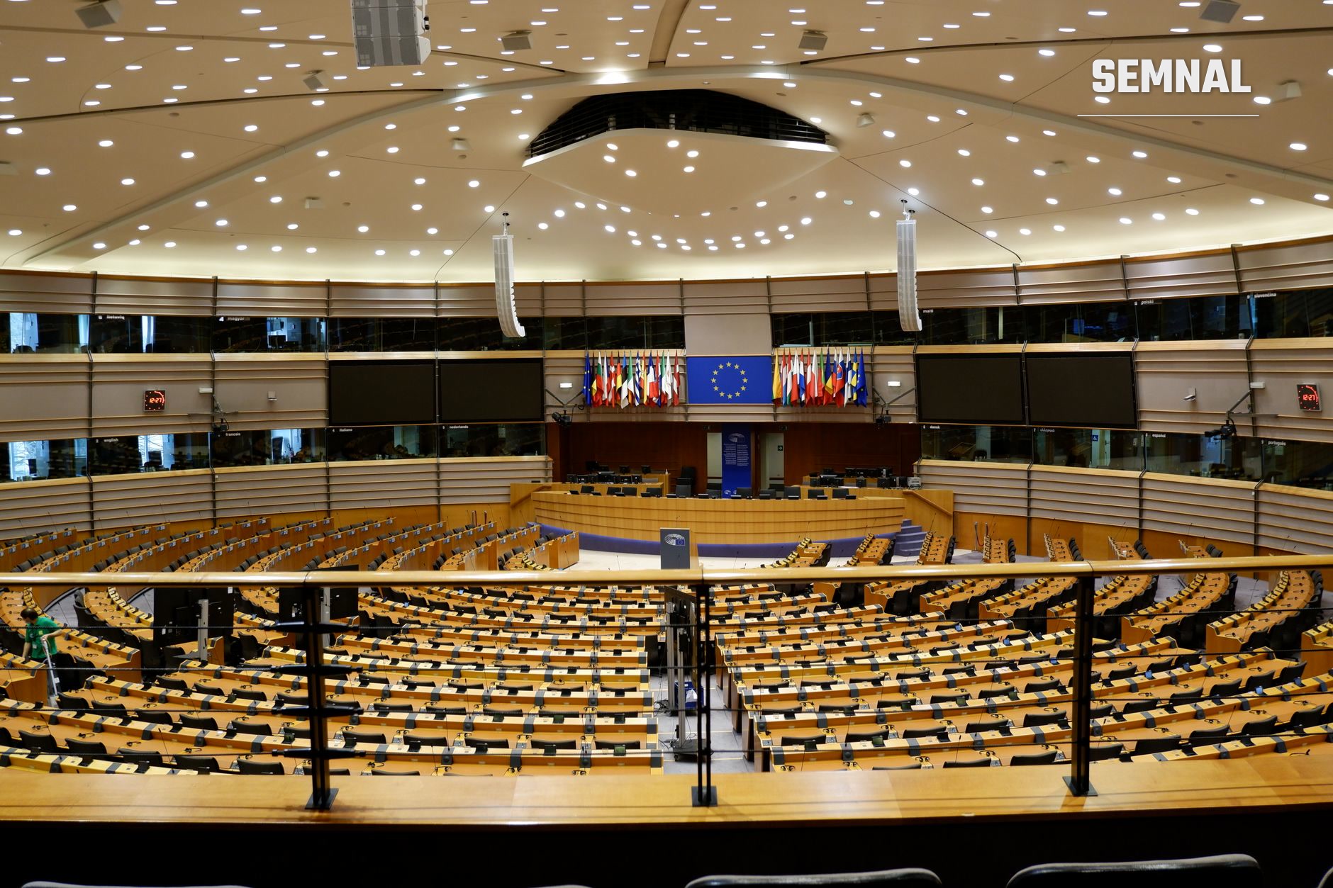Hemicyclul Parlamentului European din Bruxelles cu scaunele eurodeputaților în amfiteatru, steaguri ale statelor membre și steagul UE pe tribună, iluminat modern cu spoturi LED în plafon. (Foto: Jonas Horsch / Pexels)