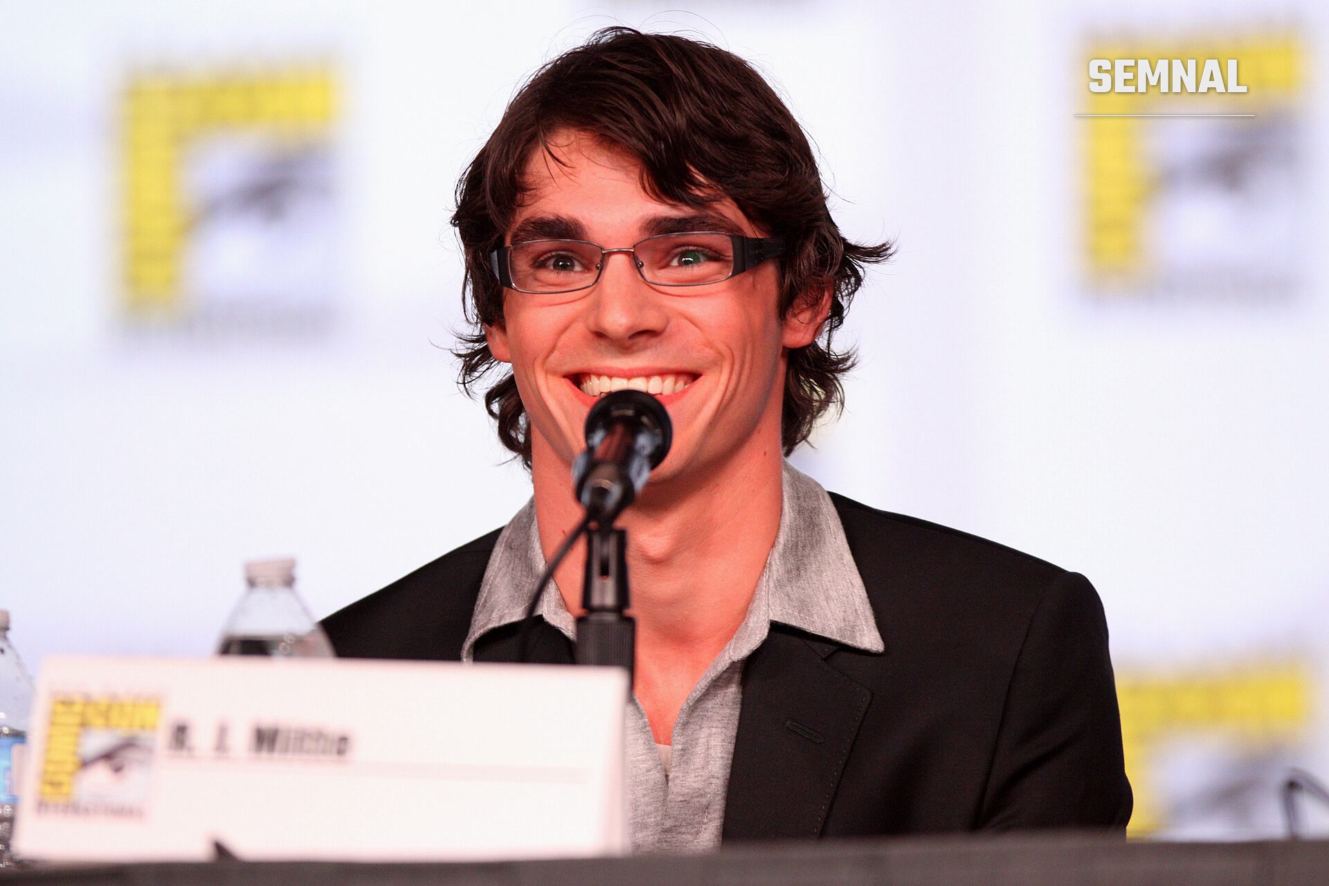 RJ Mitte zâmbind în fața unui microfon, la o masă de conferință; poartă ochelari cu ramă subțire și sacou negru peste o cămașă gri (Foto: Gage Skidmore from Peoria, AZ, United States of America)