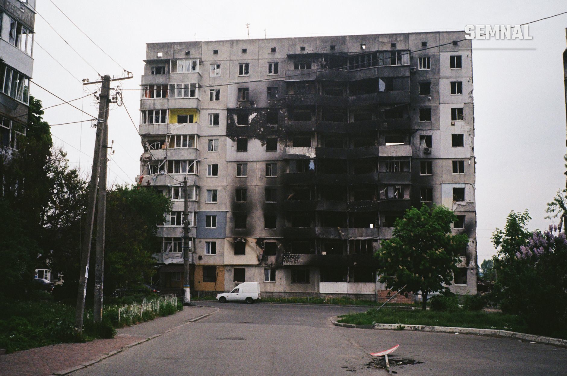 Bloc de locuințe cu nouă etaje, ars și avariat de bombardamente, cu geamuri sparte și o dubă albă pe stradă, în Ucraina. (Foto: Mykhailo Volkov / Pexels)
