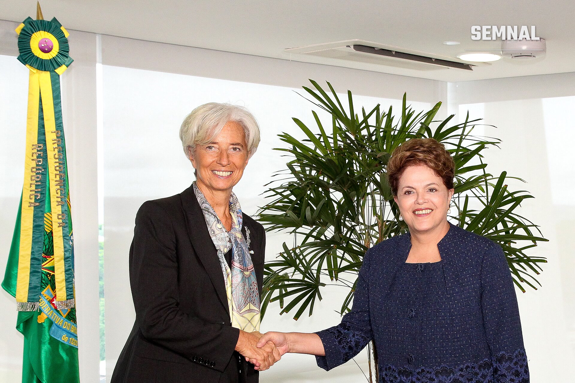 Christine Lagarde strânge mâna unei femei zâmbitoare, într-o încăpere luminoasă, lângă o plantă mare și un fanion verde-galben cu inscripția „Presidência da República”. (Foto: Dilma Rousseff from Brasil)