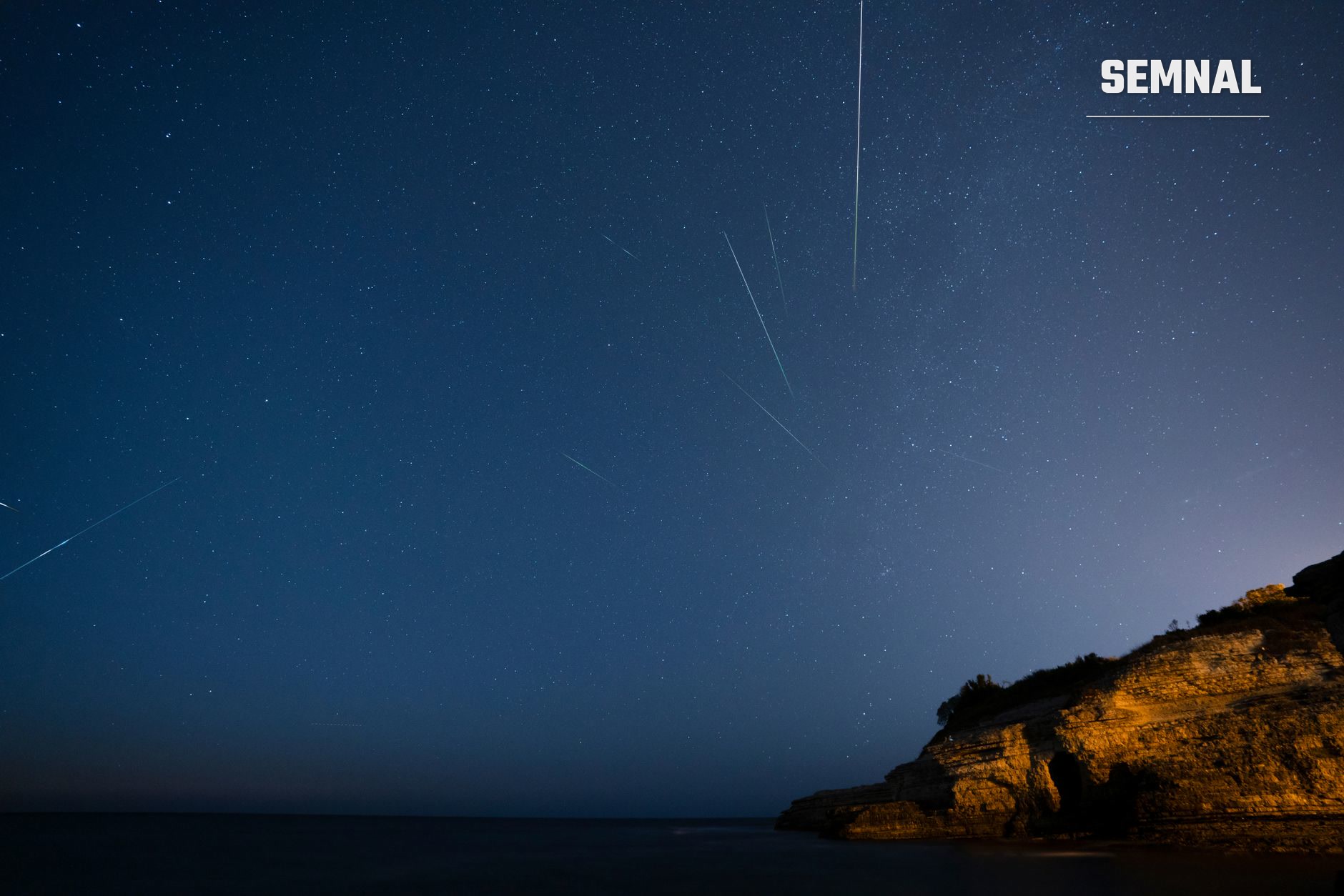 Cer nocturn înstelat, cu mai multe dâre de meteori deasupra mării și a unei stânci luminate pe malul din dreapta. (Foto: FURKAN GÜNEȘ / Pexels)