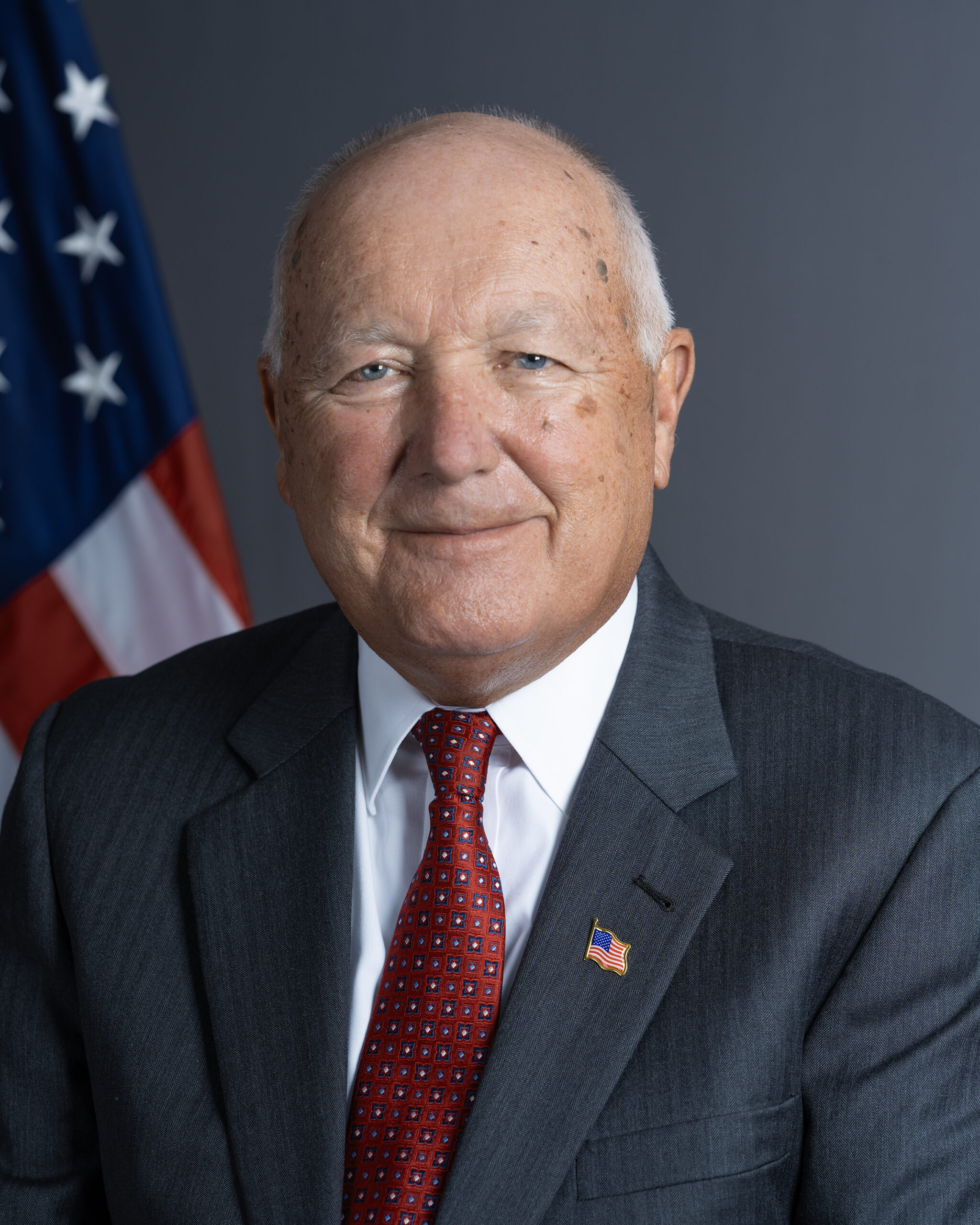 Portretul oficial al lui Pete Hoekstra, ambasadorul SUA în Canada, în costum, pe fundal albastru cu drapelul american