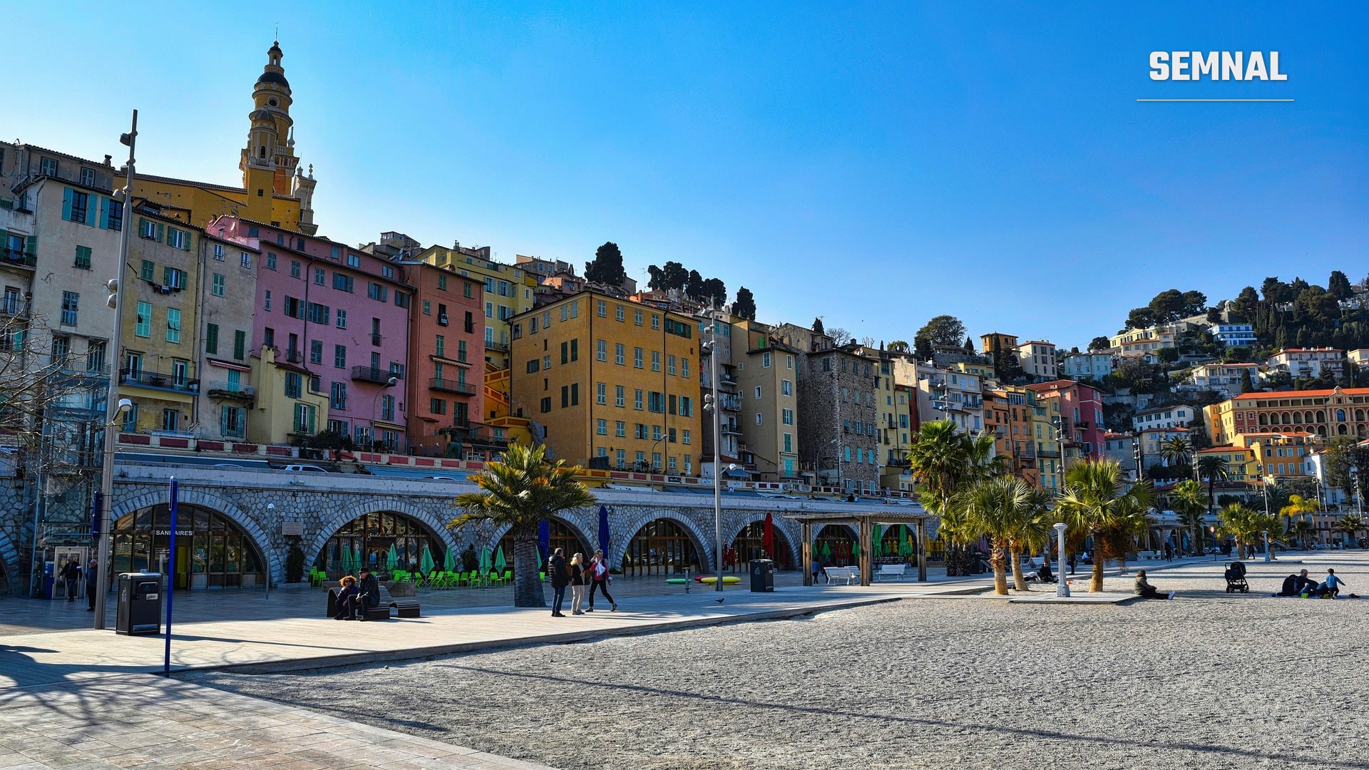 Vedere spre plaja și centrul vechi al orașului Menton, cu clădiri colorate și turnul-clopotniță al Basilicii Saint-Michel-Archange pe fundal