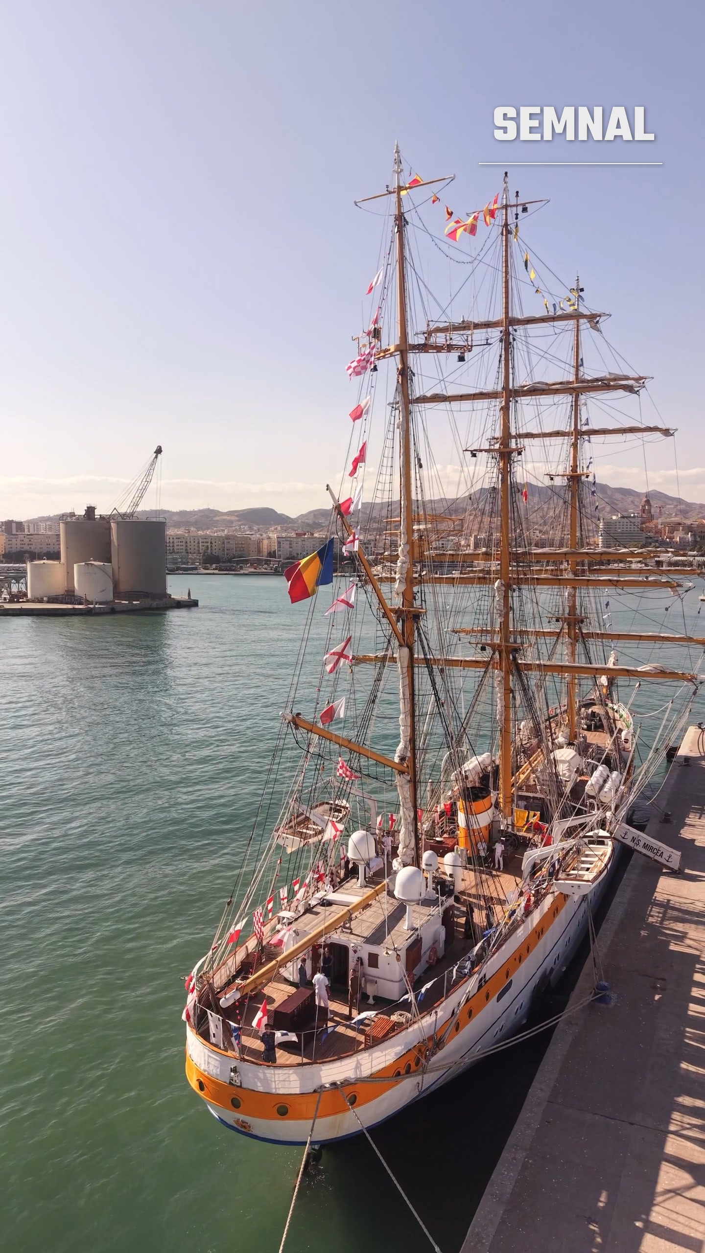 Velierul la dană, cu tricolorul la arboret și orașul Málaga în fundal