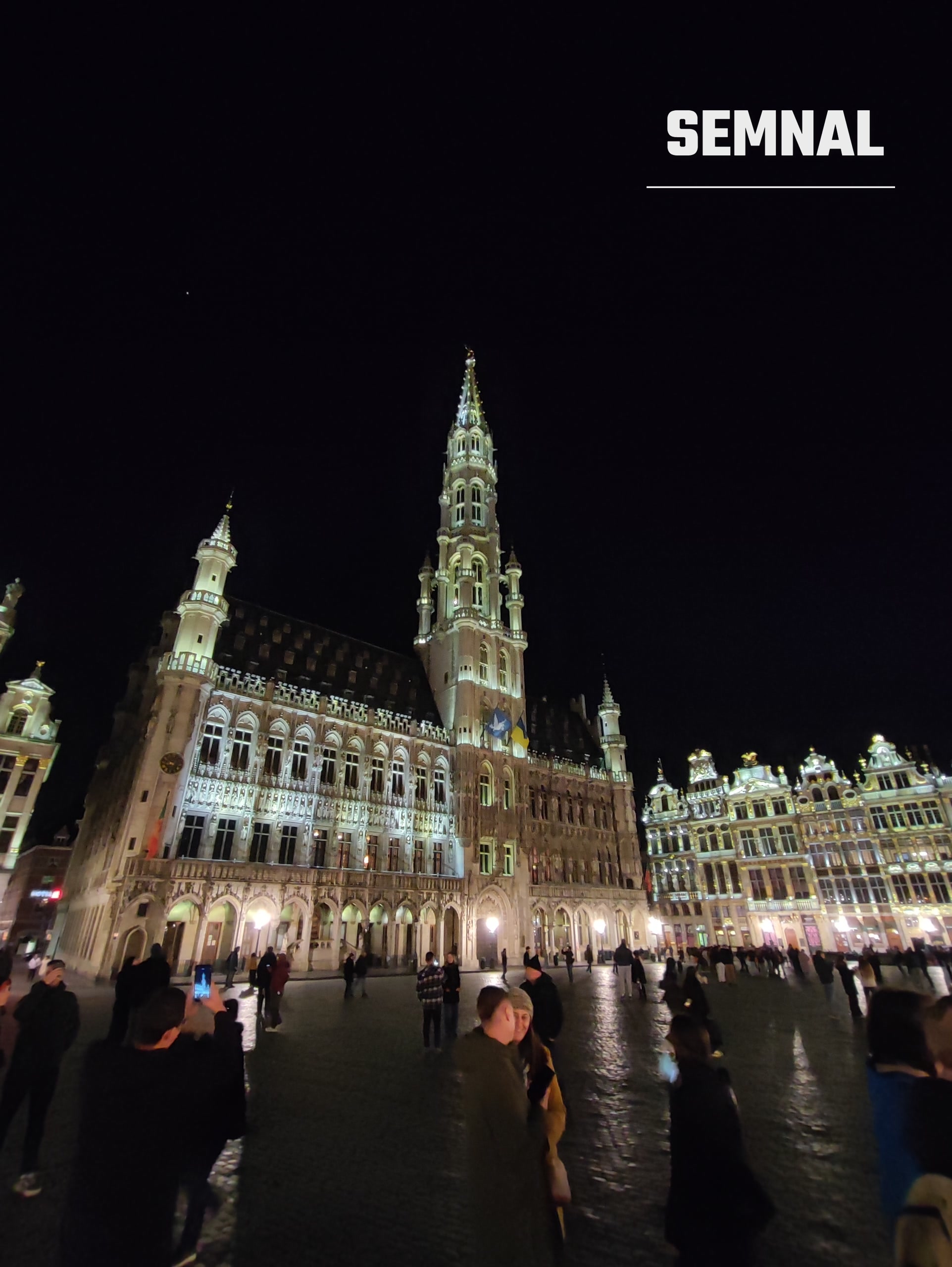 Fațadele Grand-Place, iluminate noaptea, cu vizitatori pe pavajul umed