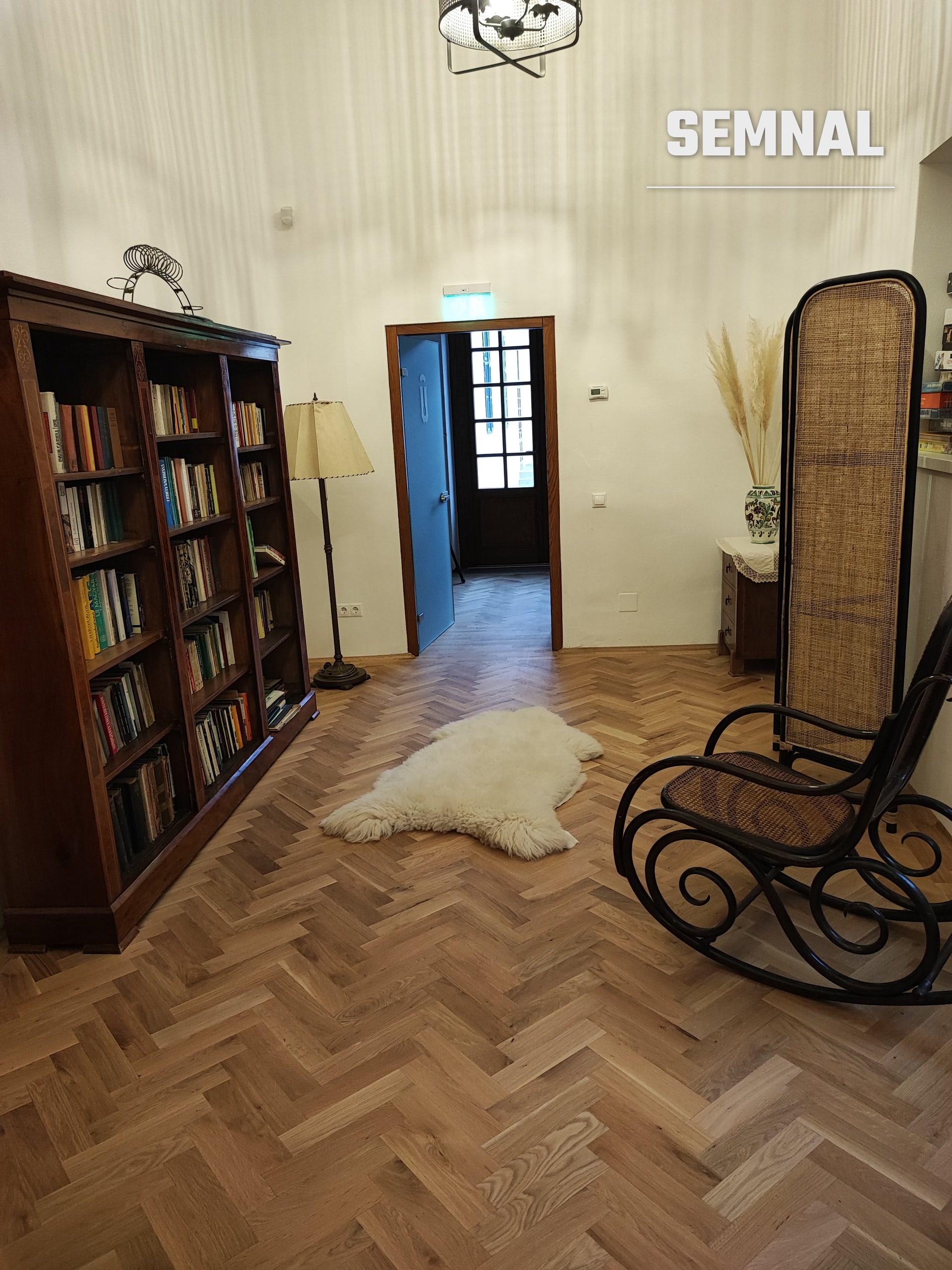 Cameră cu bibliotecă din lemn masiv, un scaun balansoar din răchită și blană albă pe podea