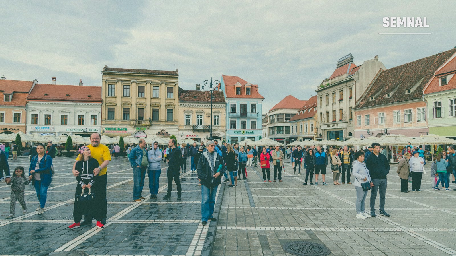 Oameni plimbându-se pe o stradă pietonală dintr-o piață istorică din Brașov, înconjurați de clădiri colorate