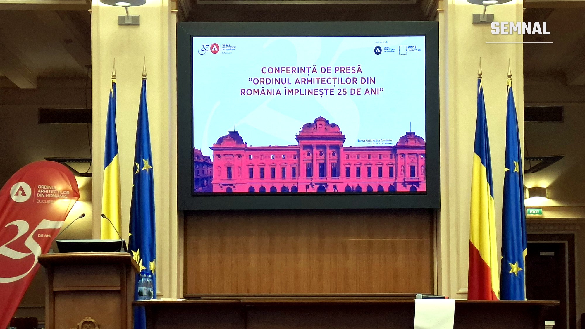 Sala conferinței de presă, cu ecranul care afișează titlul „Ordinul Arhitecților din România împlinește 25 de ani", între steagurile României și ale Uniunii Europene