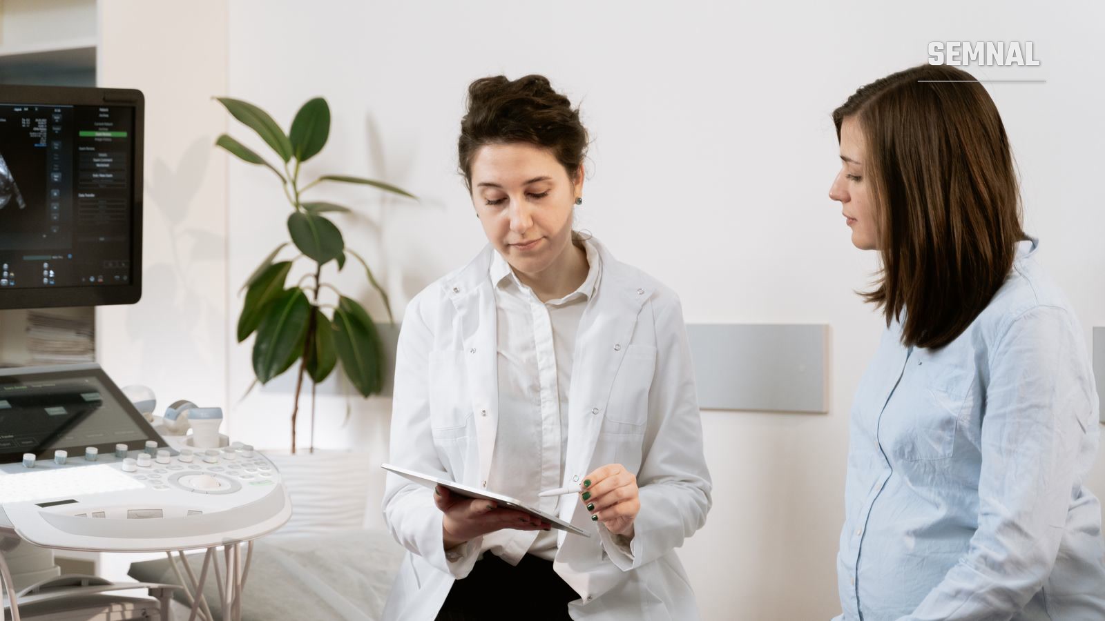 Medică în halat alb arată o tabletă unei femei însărcinate, într-un cabinet de obstetrică cu aparat de ecografie
