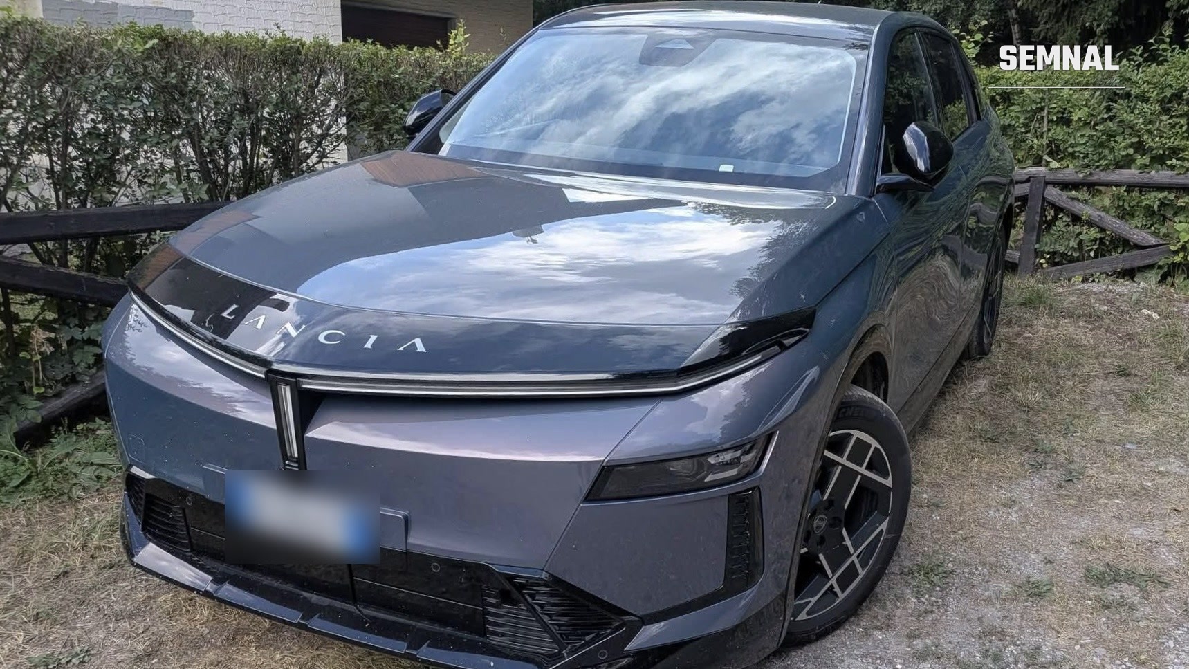 Lancia Gamma electric gri metalizat, văzut din față și din dreapta, parcat pe o alee cu pietriș, cu logo-ul Lancia pe capotă și semnătura luminoasă frontală