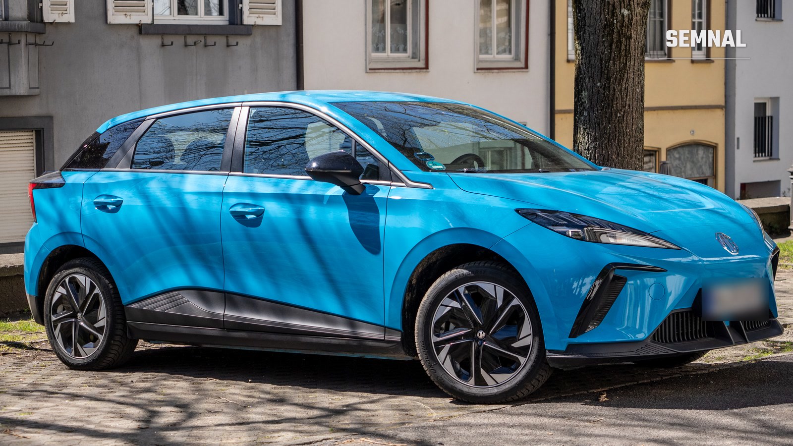 MG4 Electric albastru, văzut din lateral și din față, parcat pe o stradă pavată, în fața unor clădiri vechi