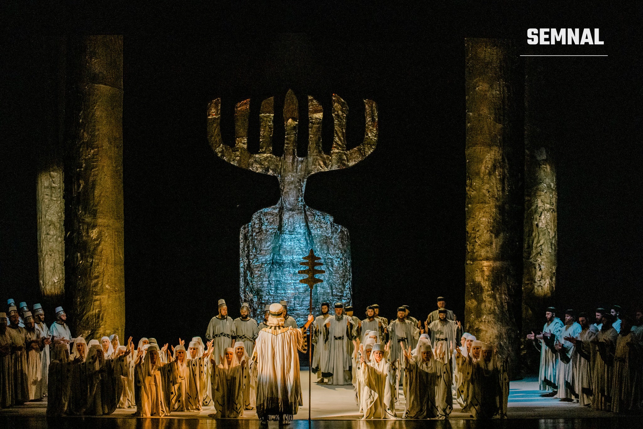 Scenă din spectacolul „Nabucco” la Opera Națională București, cu coriști în costume albe, cu brațele ridicate, în fața unui decor în formă de sfeșnic