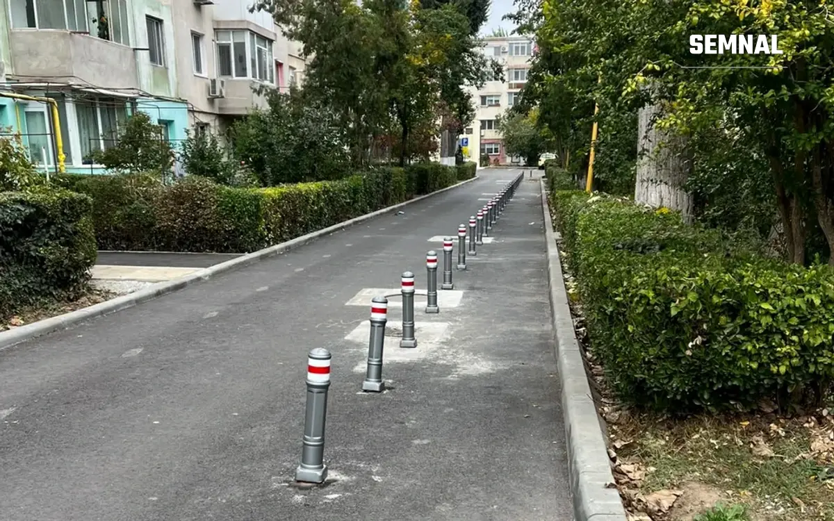 Fâșie de asfalt între două garduri vii, cu un șir de stâlpișori gri cu benzi roșii, lângă blocuri de locuințe din Constanța
