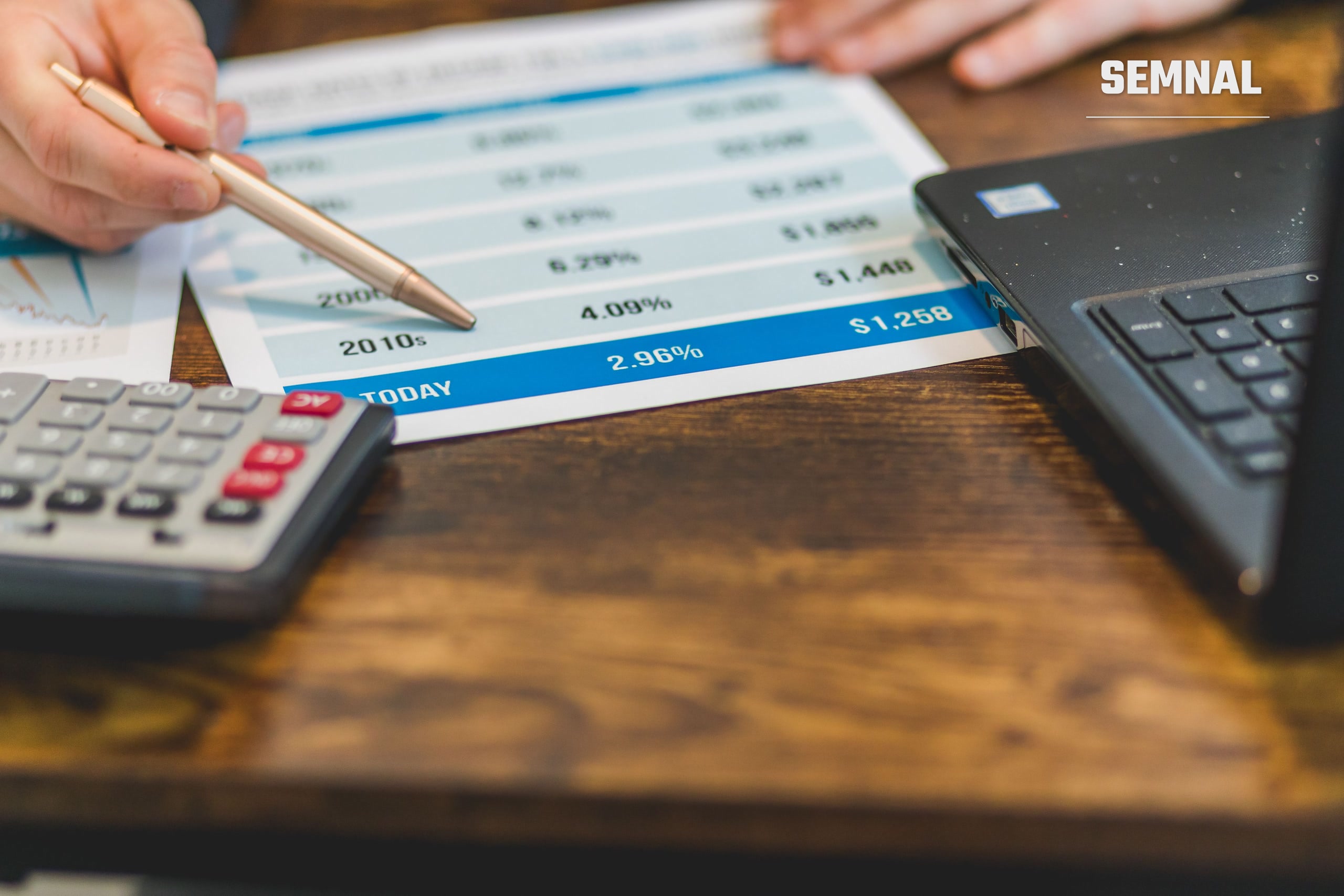 Mână cu pix care analizează un document cu cifre, lângă un calculator și un laptop pe o masă, fotografie ilustrativă