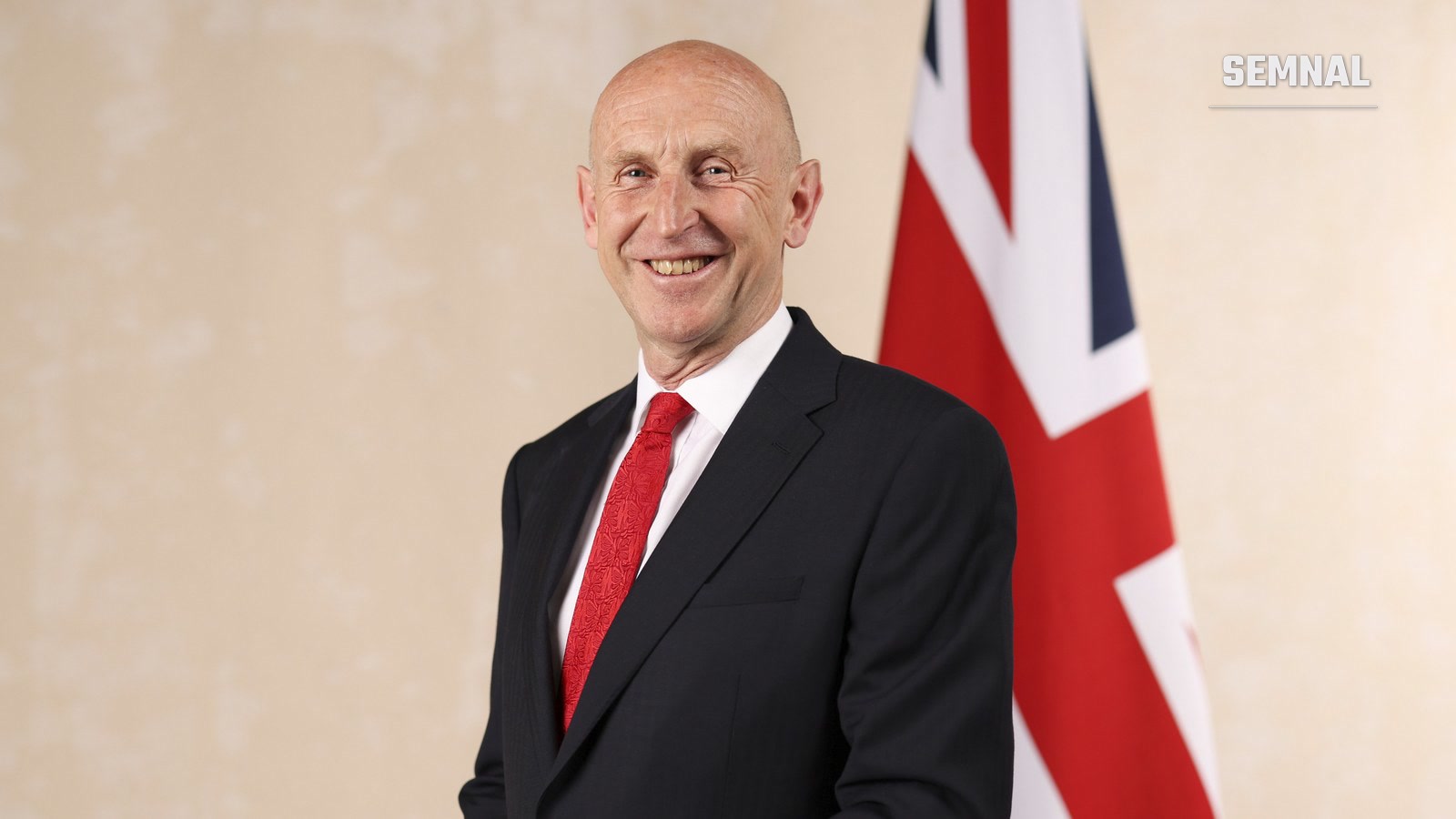 John Healey, cancelarul Marii Britanii, zâmbind, în costum închis la culoare și cu cravată roșie, lângă un drapel britanic.